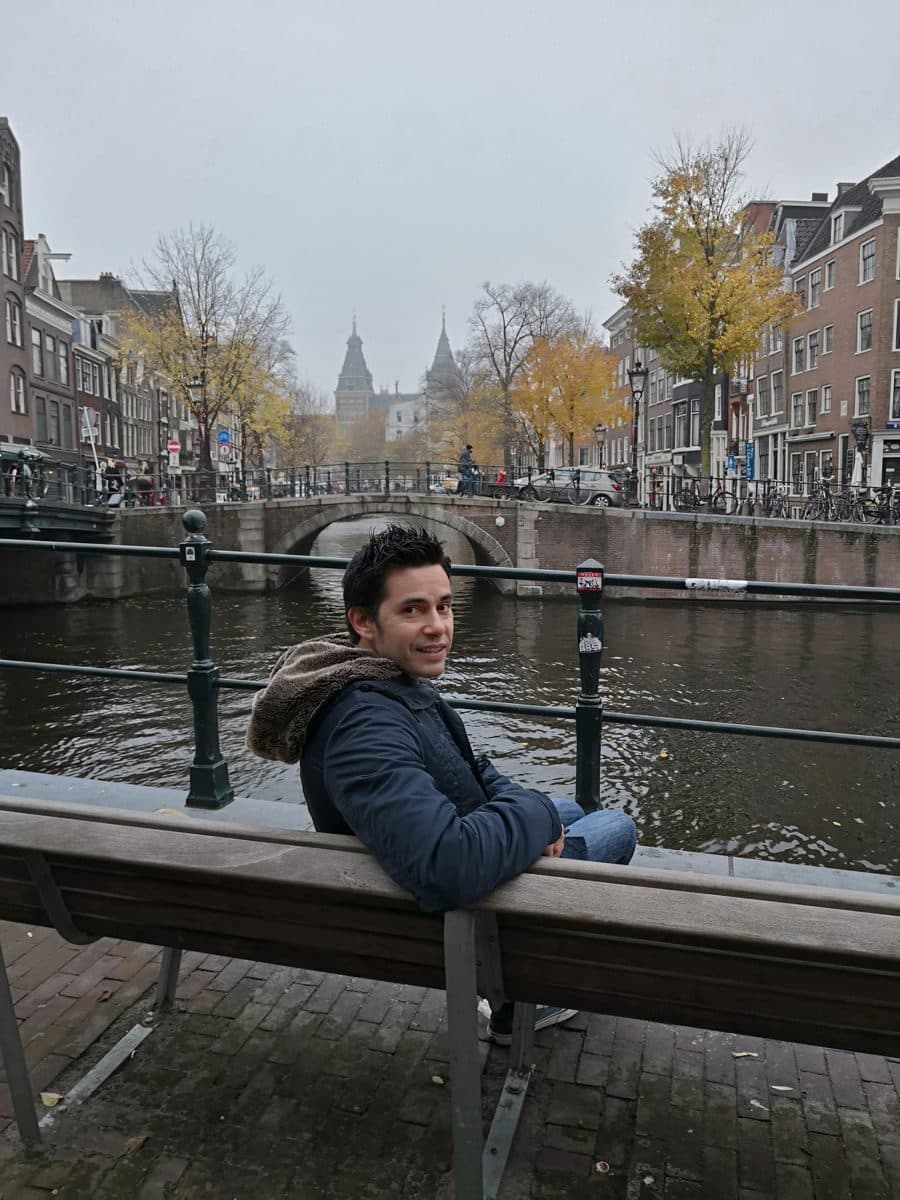 Diego Fresno sitting on a bench beside an Amsterdam canal under a completely overcast sky, with yellow autumn leaves still on the trees and a stone bridge behind him