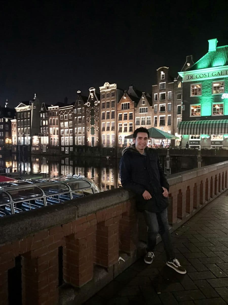 Diego Fresno leaning on a railing on the Damrak in Amsterdam at night, with the lit canal houses across the water