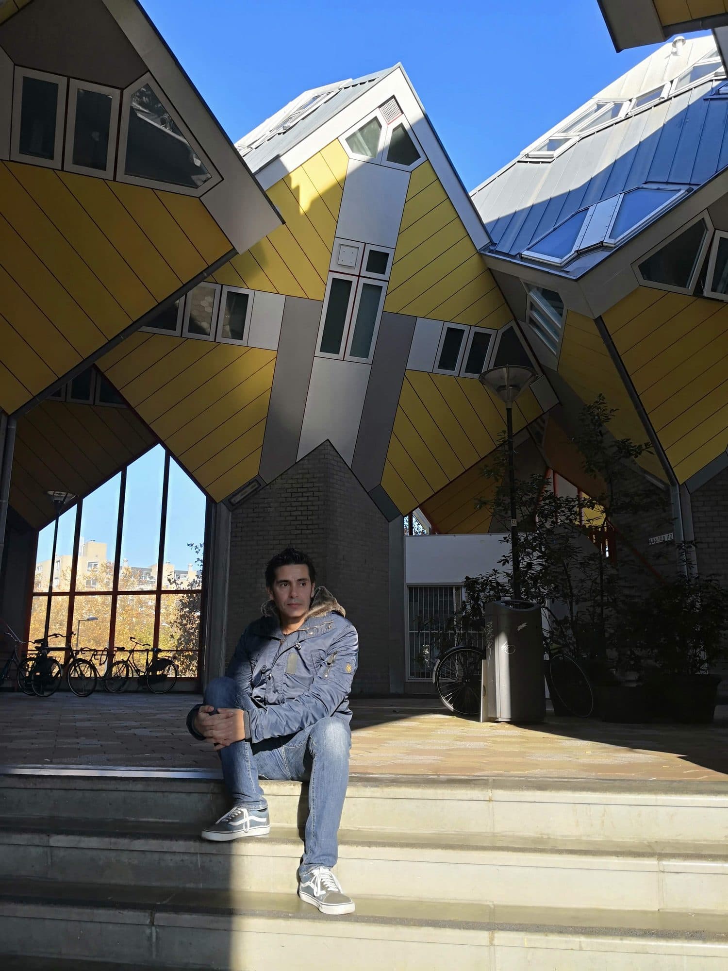 Diego Fresno sitting on steps beneath the yellow cube houses in Rotterdam, tilted on their corners against a cloudless blue sky, with bicycles parked under the arch behind him