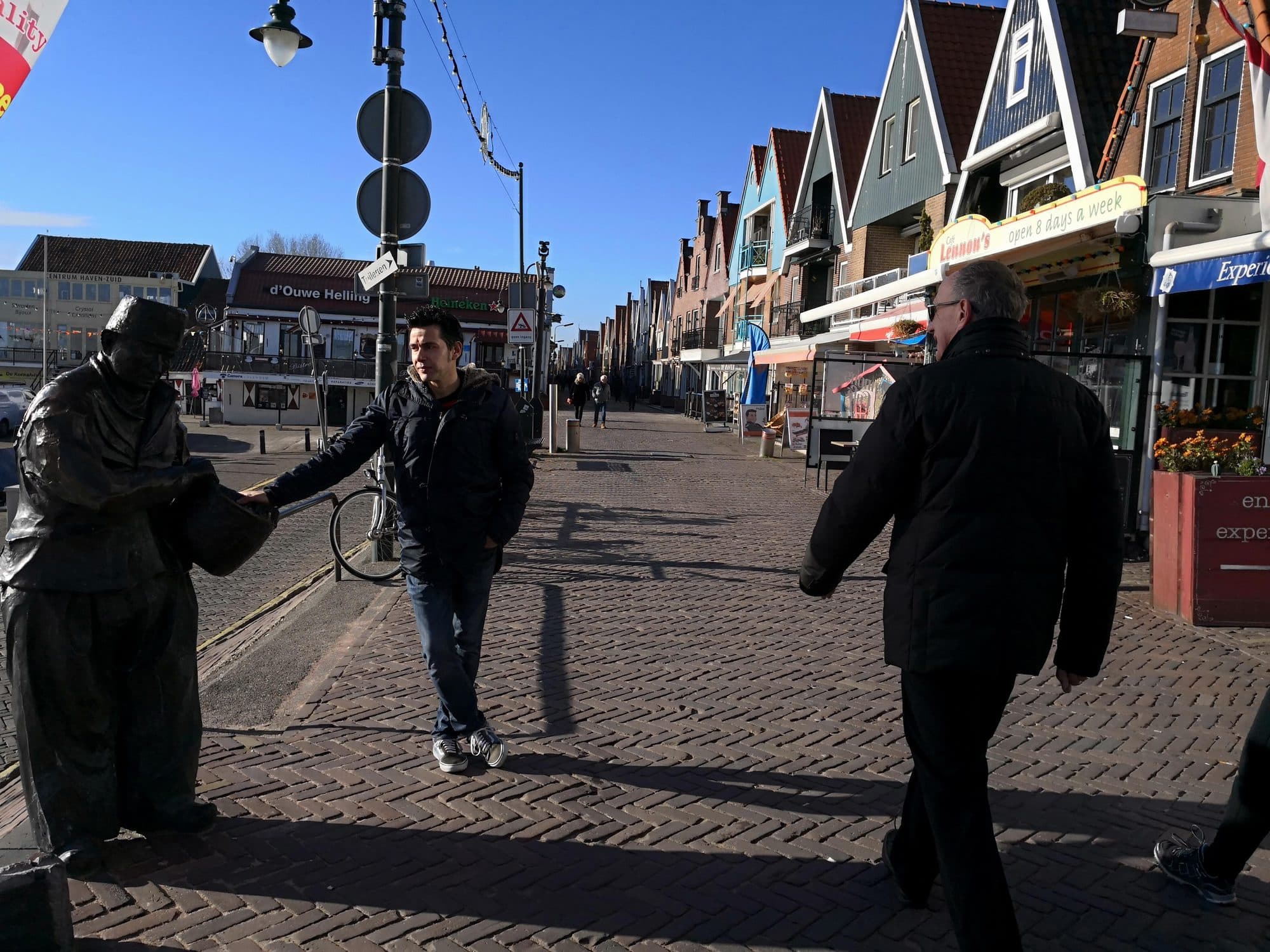 The Haven in Volendam almost empty at midday, with the row of painted gabled shopfronts along the harbour street and a bronze statue of a fisherman in the foreground, under a clear winter sky