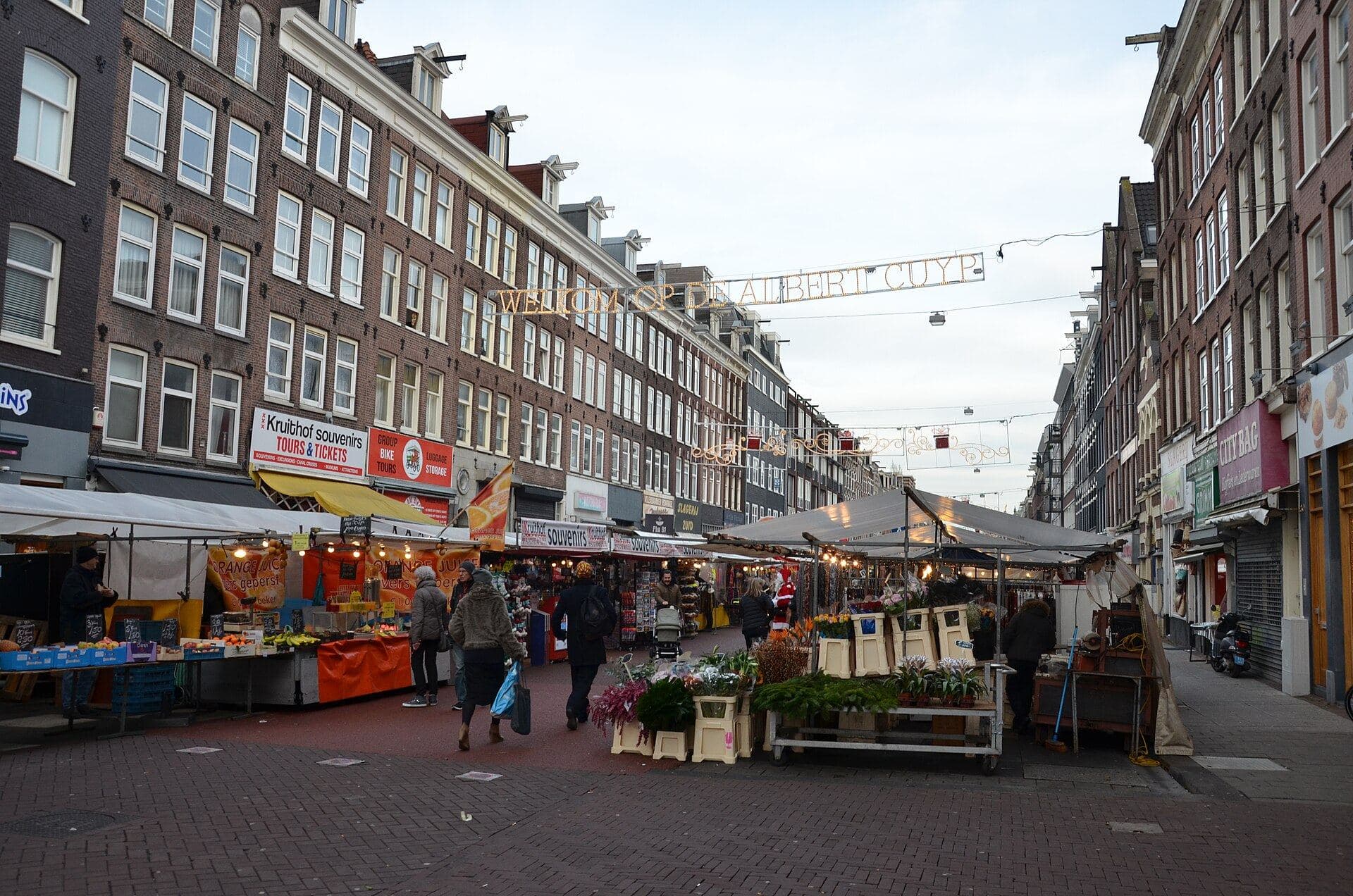 The entrance to the Albert Cuyp market in Amsterdam under an illuminated sign reading Welkom op de Albert Cuyp, with covered stalls selling fresh orange juice and flowers along a brick street on a grey winter day