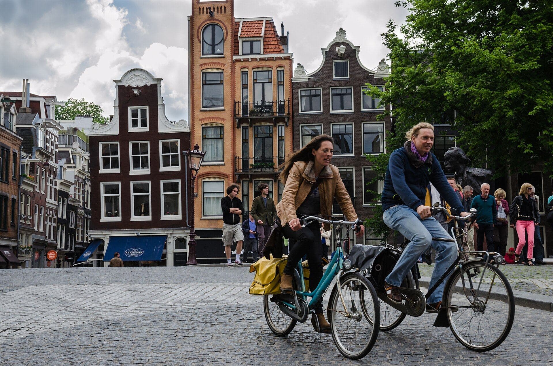 Two people cycling across a cobbled bridge in central Amsterdam on upright city bikes with panniers, neither wearing a helmet, with canal houses and stepped gables behind them