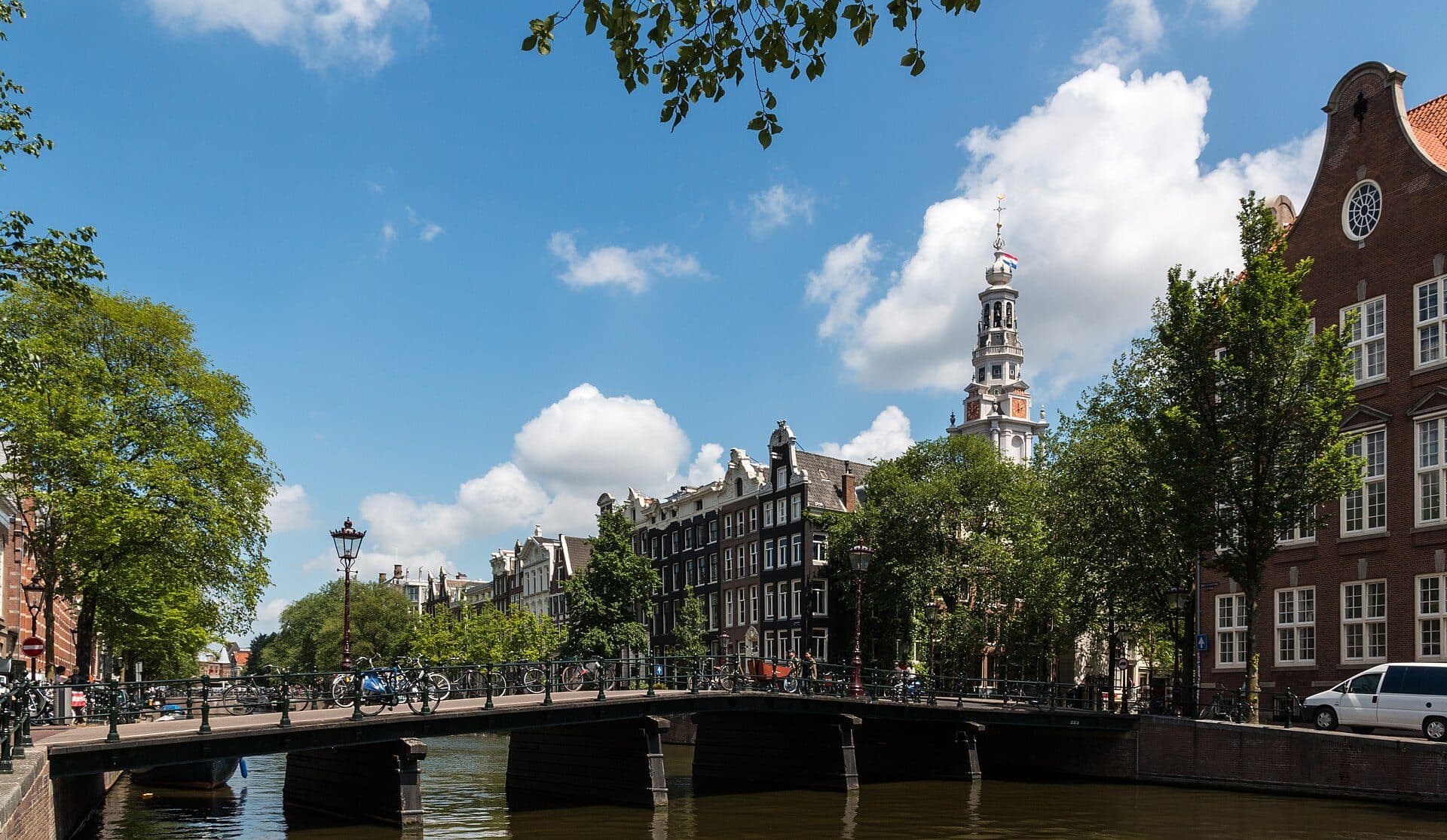 A canal bridge in Amsterdam lined with parked bicycles on a clear summer day, with the tower of the Zuiderkerk rising behind the trees