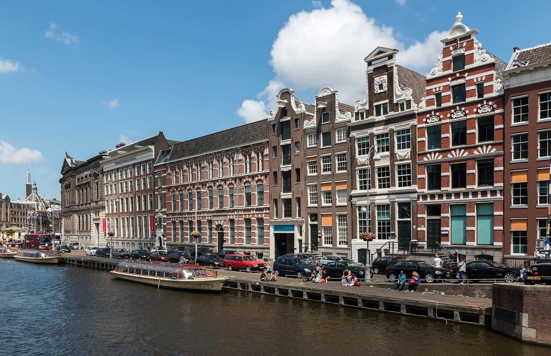 Canal houses with stepped and bell gables along the Rokin in central Amsterdam, with a glass-roofed tour boat moored below and people sitting on the quayside