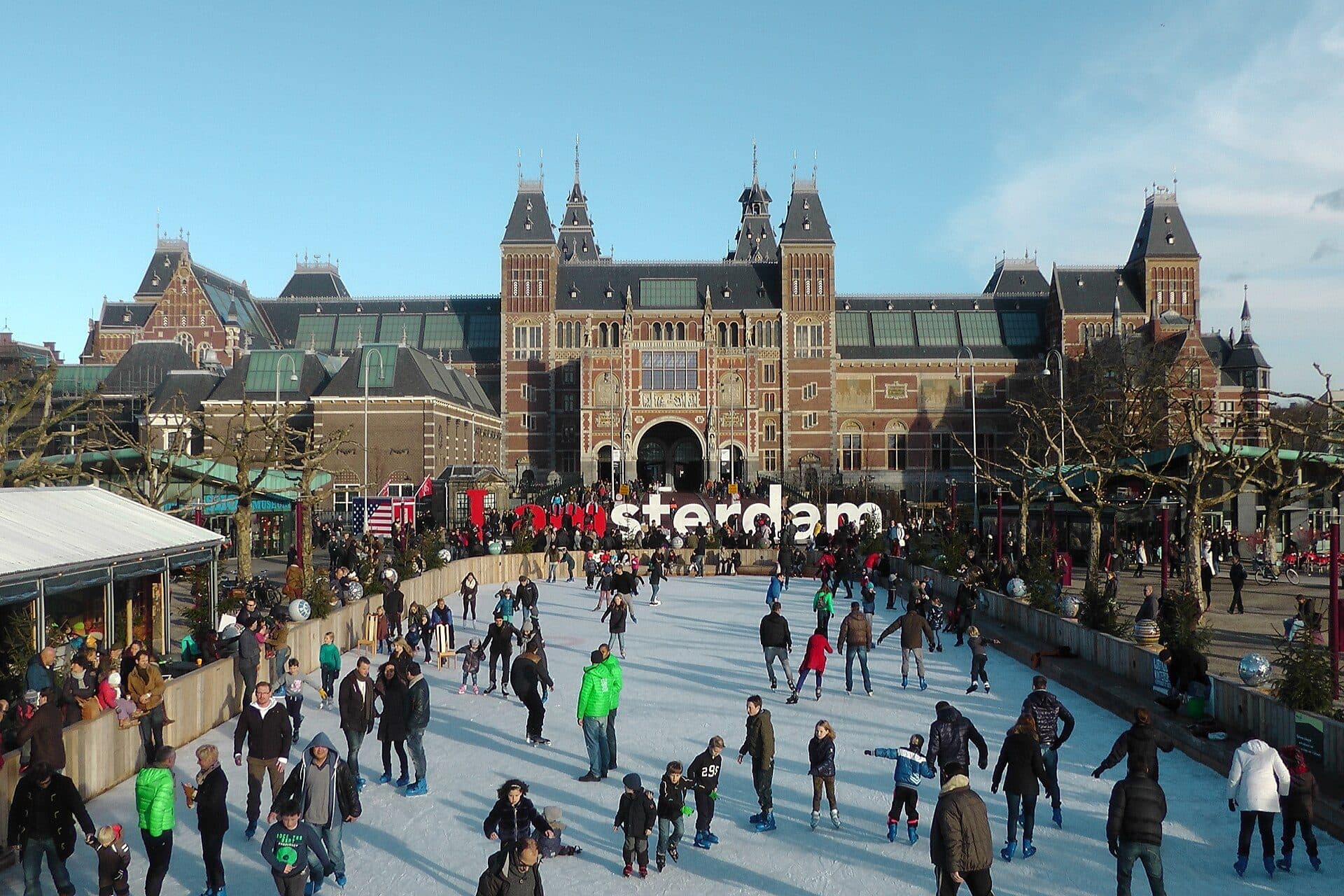 The outdoor ice rink on Museumplein crowded with skaters on a clear winter day, with the Rijksmuseum behind it, the I amsterdam letters on the far side and the wooden market huts of the Christmas Village along the left edge
