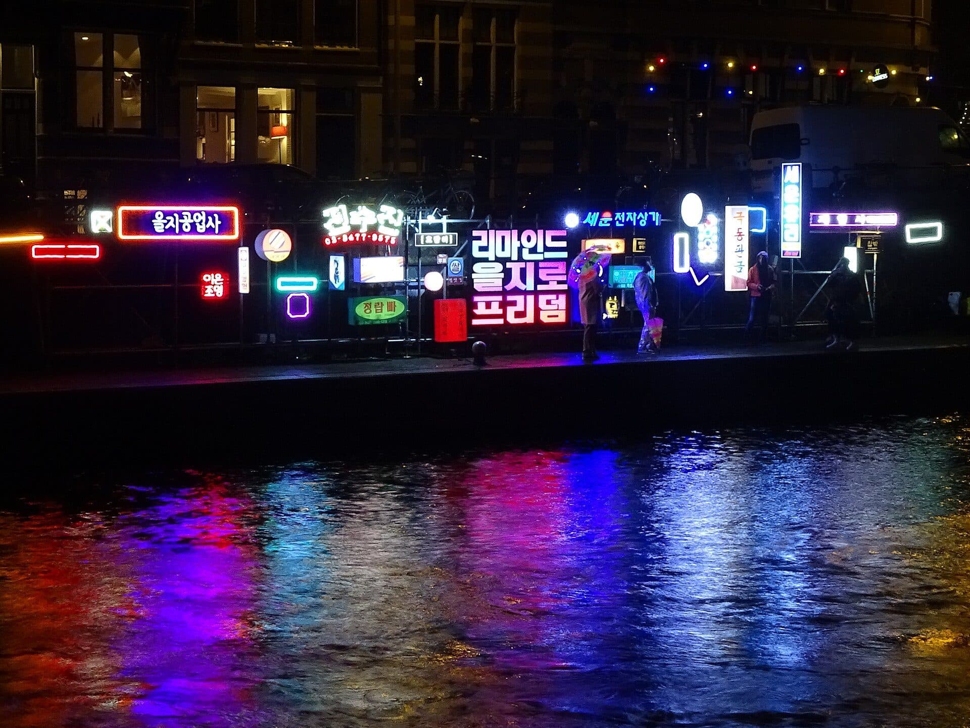 A wall of coloured neon signs in Korean lettering installed along the Schippersgracht during Amsterdam Light Festival, reflected in the black canal water, with four people watching from the quayside