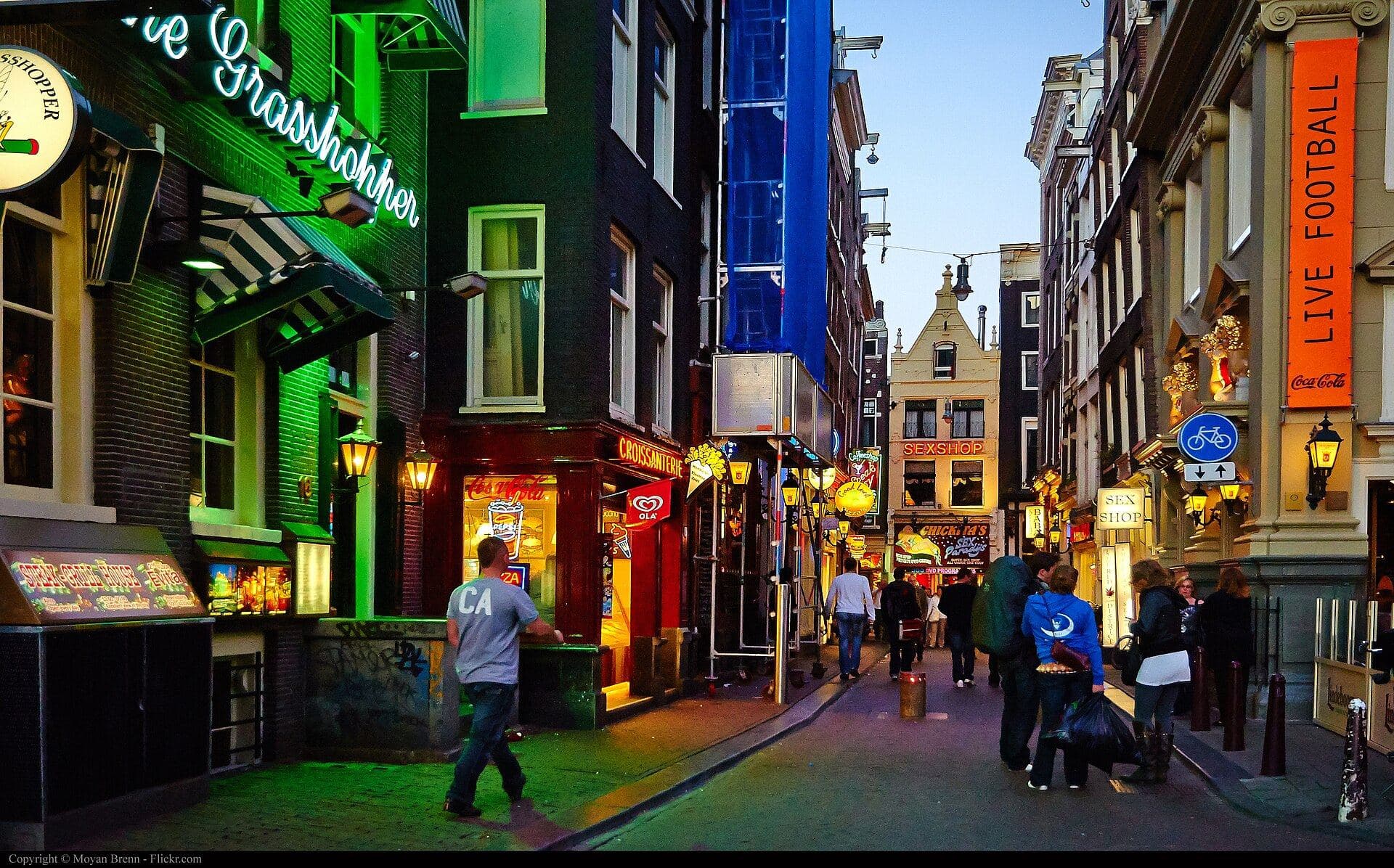 A narrow street in central Amsterdam at dusk, lit by the green neon of a coffeeshop sign, with sex shop and live football signs further along and people walking between them