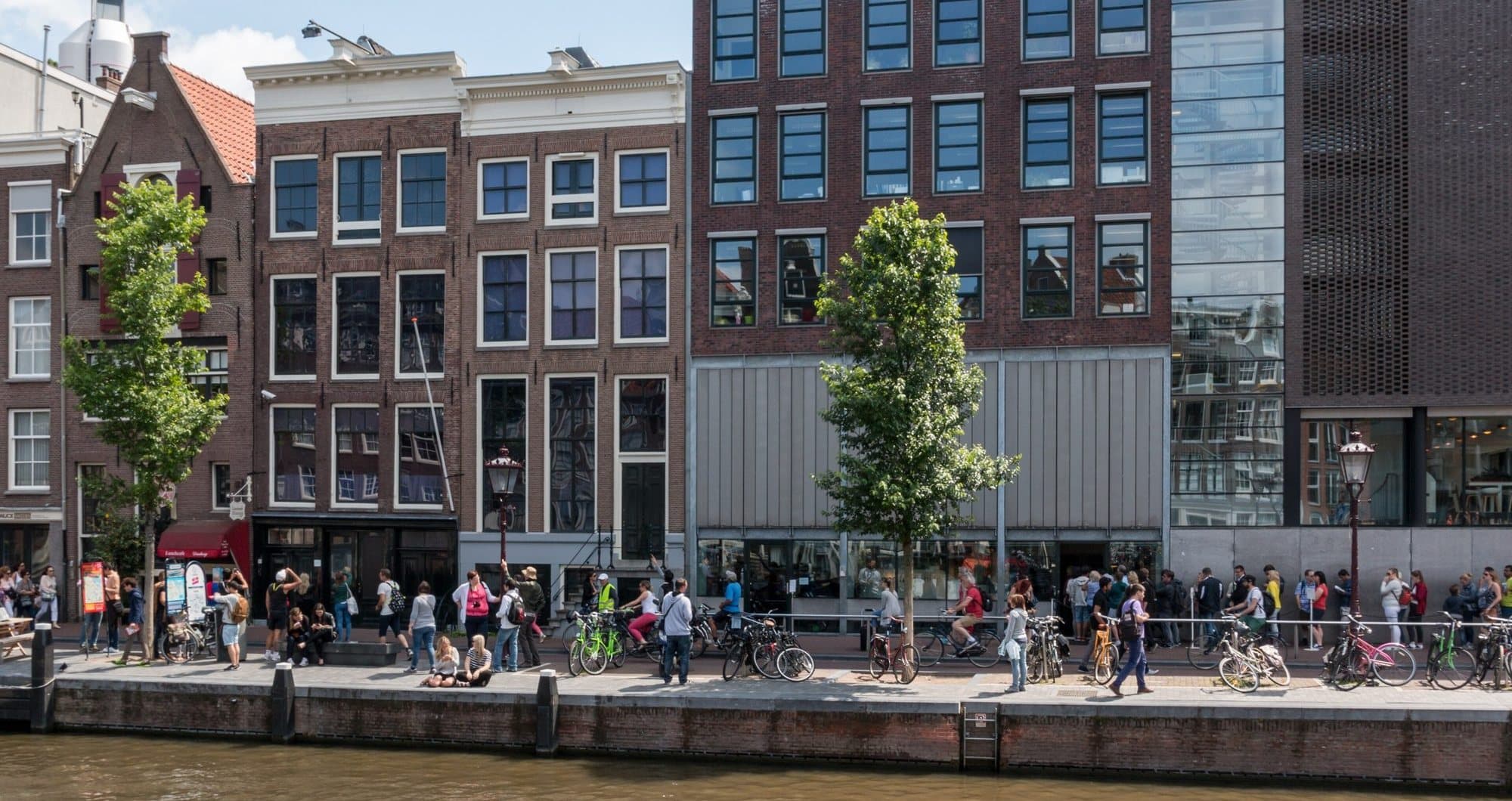 The Anne Frank House seen from across the Prinsengracht, with the original brick house beside the museum's newer entrance building and a queue of visitors running the length of the quayside