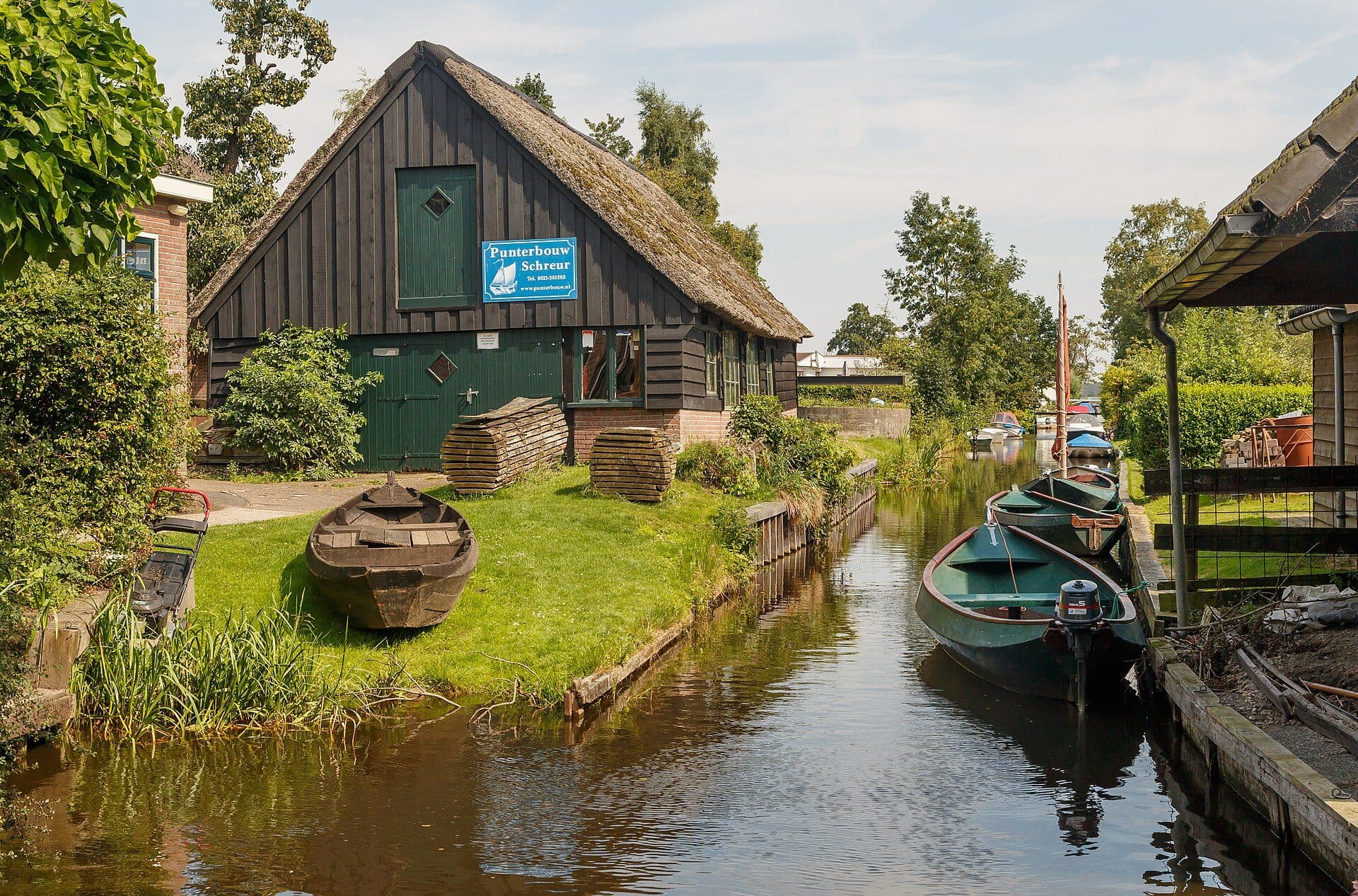 A canal in Giethoorn with a thatched farmhouse on the left bank, a wooden punt pulled up on the grass beside it and two moored green boats on the right, under summer trees