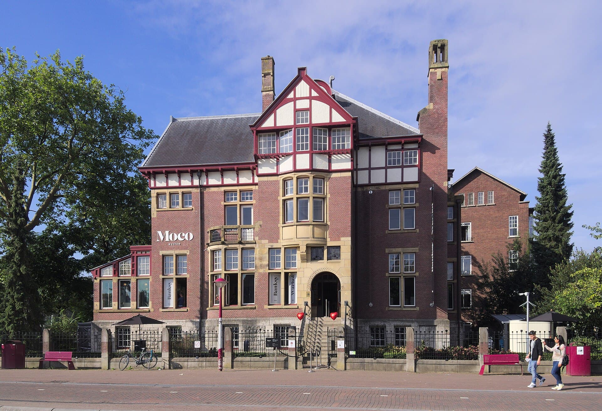 The Moco Museum in Amsterdam, a red brick and timbered 1904 villa on the Museumplein with the Moco wordmark on its side wall, Banksy and Warhol banners in the windows and pink benches along the pavement