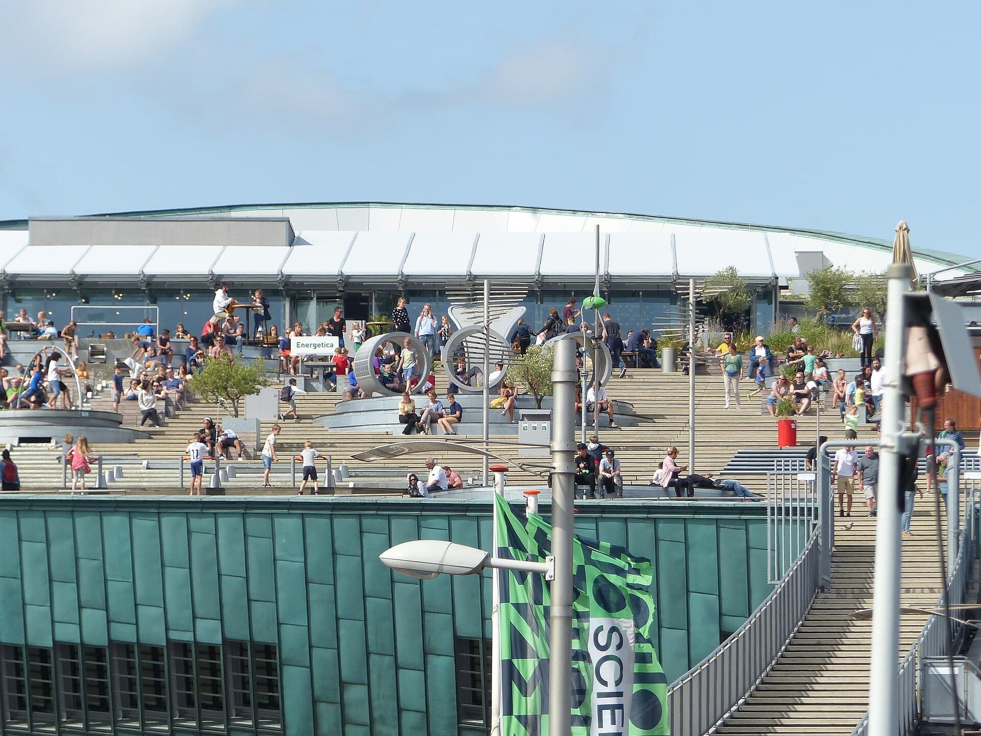The stepped roof terrace of NEMO Science Museum in Amsterdam on a sunny day, crowded with people sitting on the wide steps and at tables, with the green copper hull of the building below and the open staircase on the right
