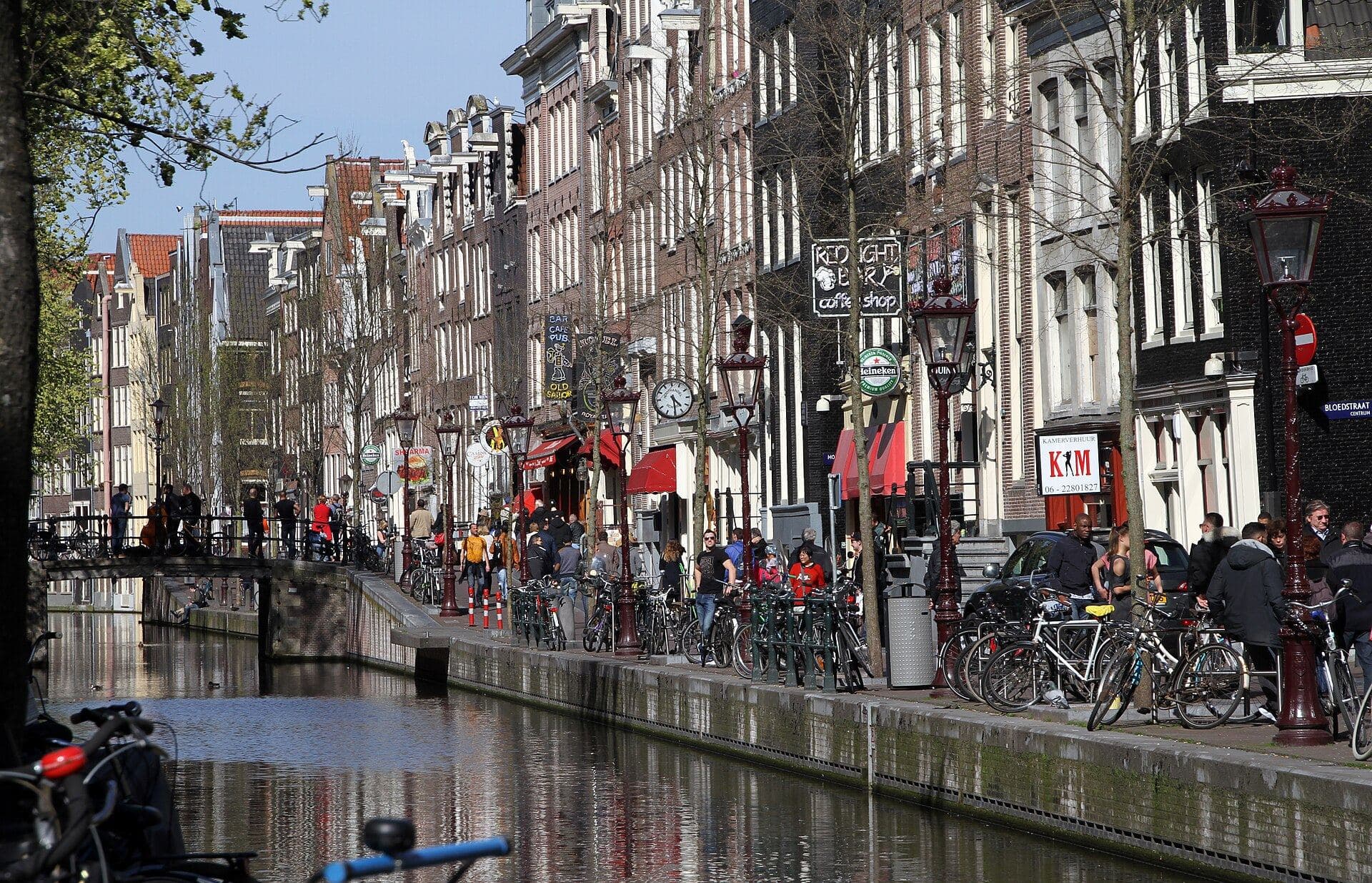 The Oudezijds Achterburgwal canal in Amsterdam's Red Light District on a sunny day, with bars and a coffeeshop sign along the quay, parked bicycles, red street lamps and people walking by the water