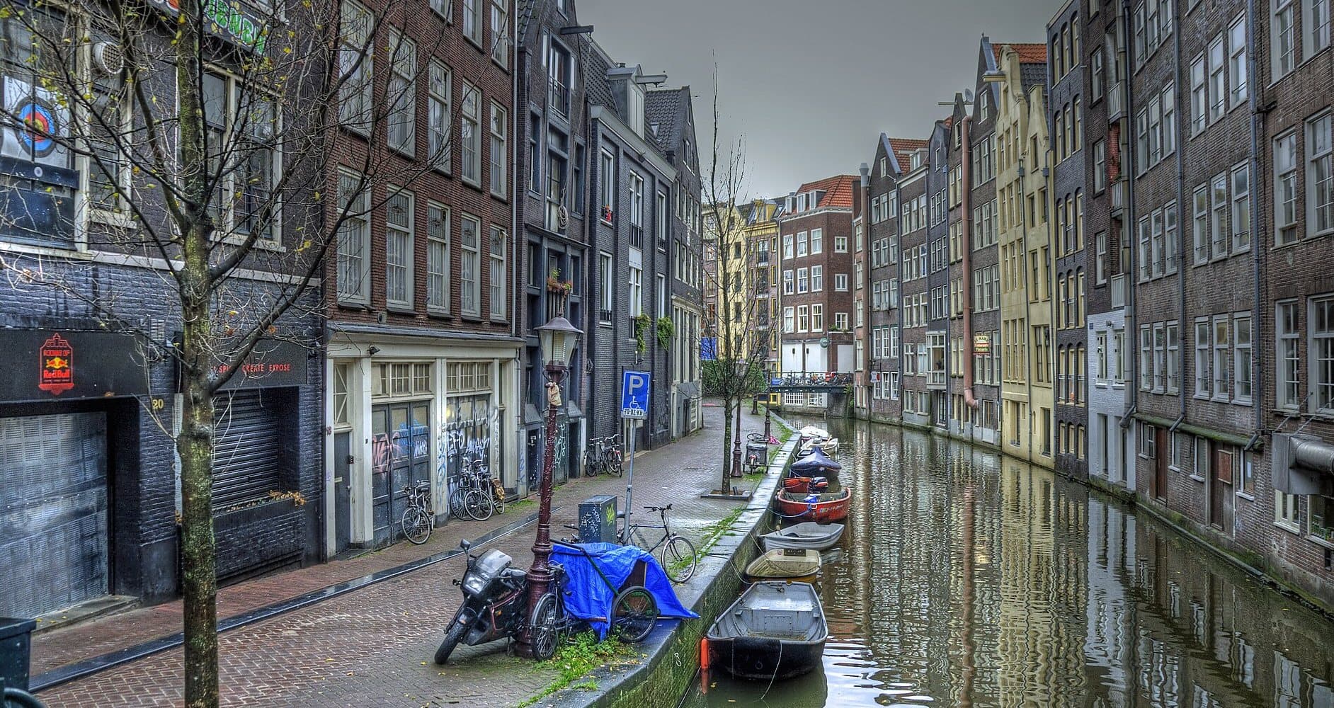 The Oudezijds Voorburgwal canal in De Wallen on a grey morning, with narrow brick houses on both banks, moored rowing boats and shuttered doorways along the quay