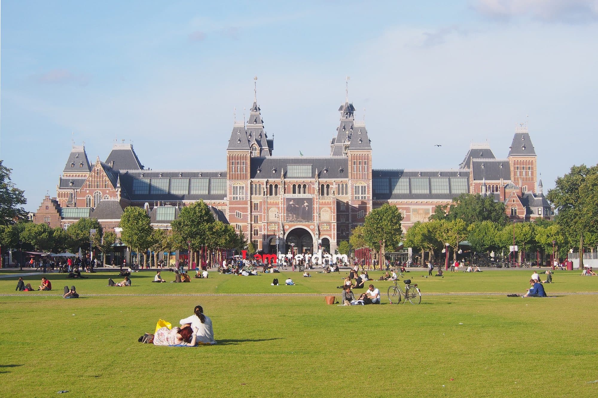 The Rijksmuseum seen across the lawn of the Museumplein on a clear day, with people sitting on the grass and the I amsterdam letters standing in front of the entrance