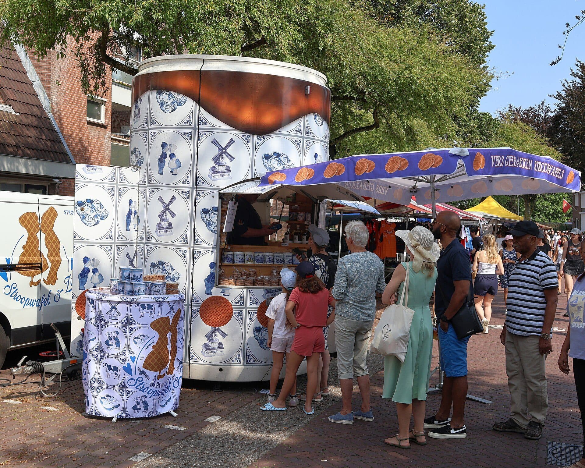 A stroopwafel stall at a Dutch street market, decorated in Delft blue tiles, with a queue of people waiting to be served
