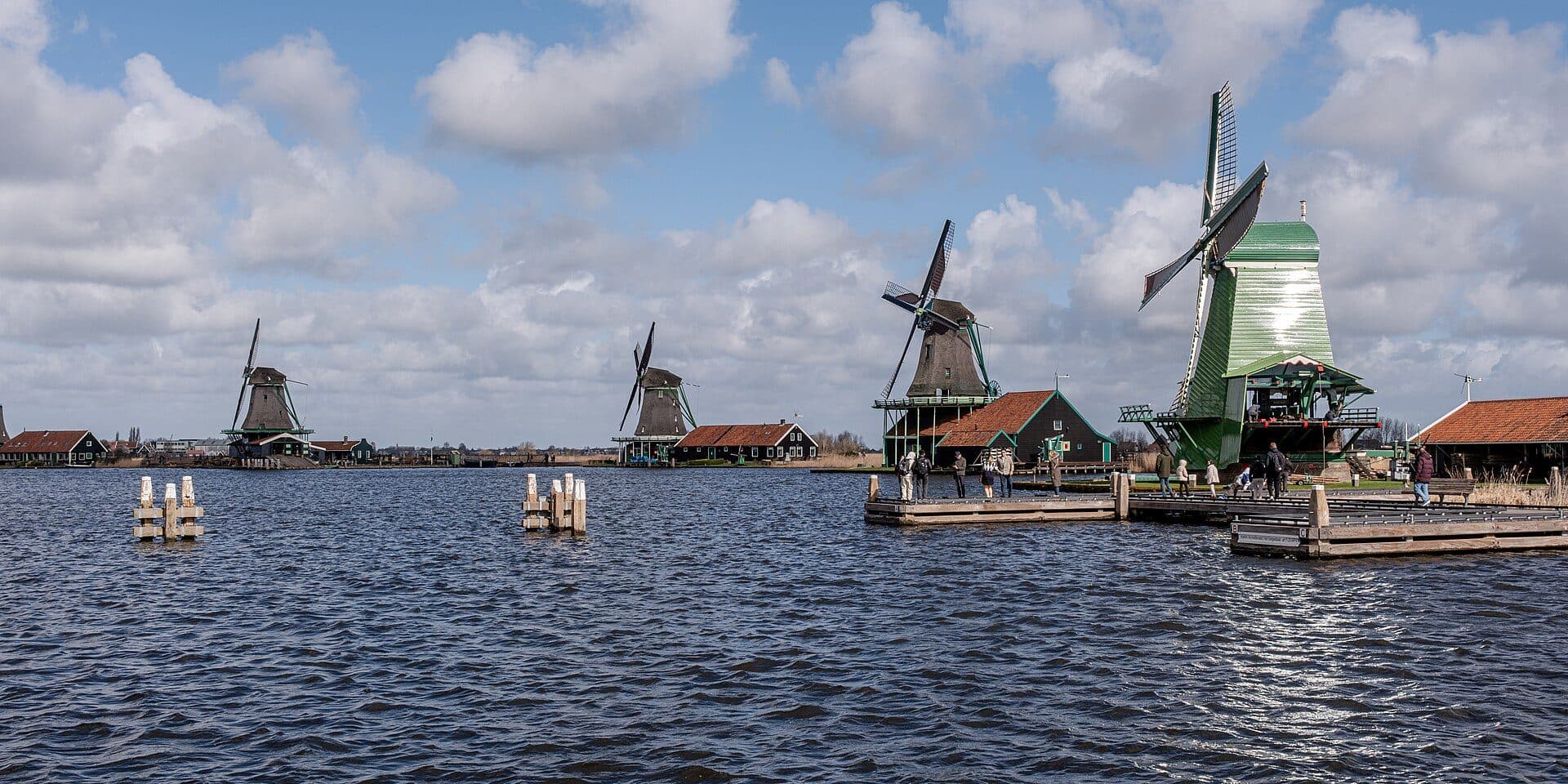Four working windmills along the water at Zaanse Schans under a sky of broken cloud, with visitors on the wooden jetty in front